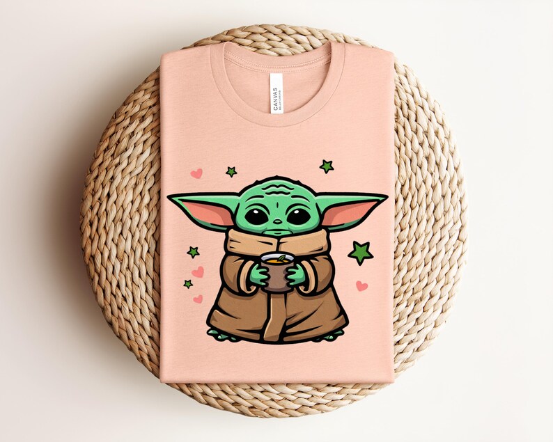 May include: A light peach-colored t-shirt featuring a cartoon illustration of a green alien character holding a cup. The character is wearing a brown cloak and has large ears. Pink hearts and green stars surround the character.