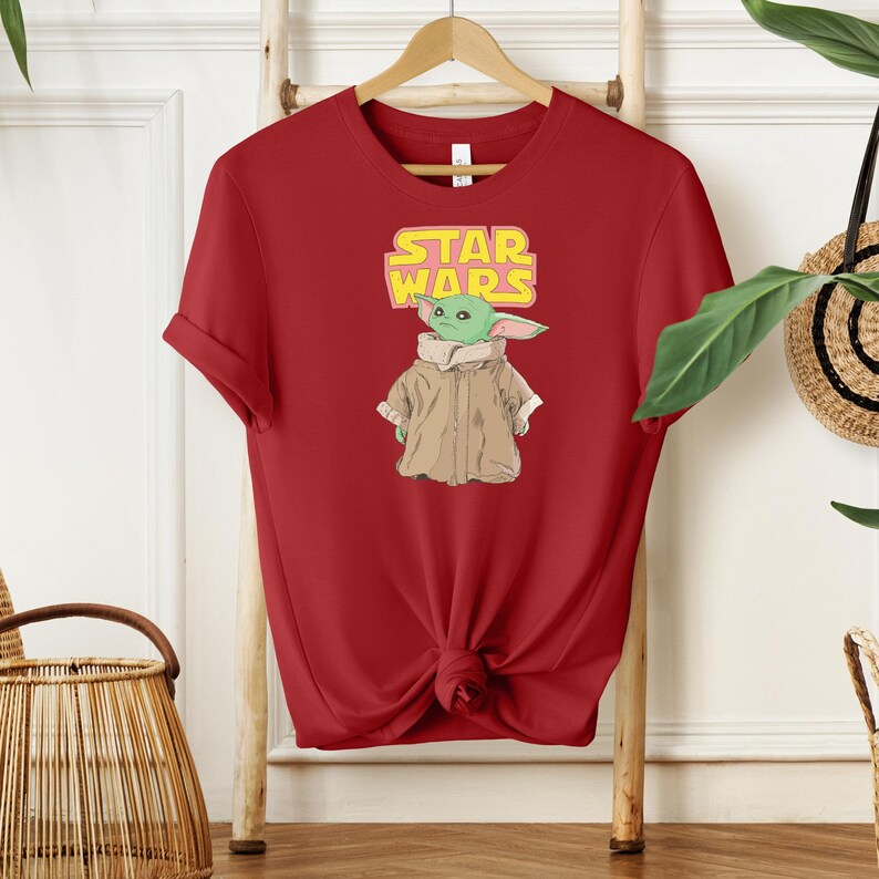 Puede incluir: Camiseta roja con un dise&ntilde;o de Grogu, el personaje de Baby Yoda de Star Wars. El dise&ntilde;o incluye el logo de Star Wars en amarillo y el personaje con un abrigo marr&oacute;n. La camiseta est&aacute; colgada en una percha de madera.