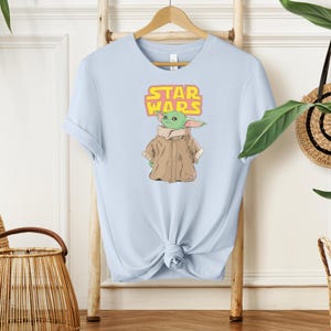Puede incluir: Camiseta azul claro con un gr&aacute;fico de un personaje alien&iacute;gena verde de Star Wars, que lleva un abrigo marr&oacute;n. El logotipo de Star Wars est&aacute; encima del personaje en amarillo y rosa. La camiseta se muestra en una percha de madera.