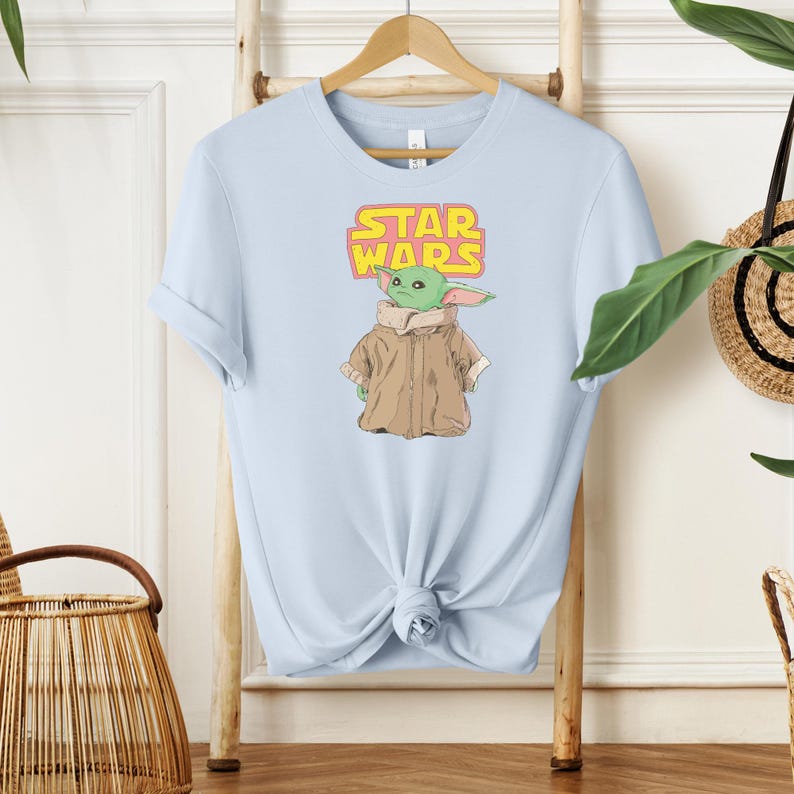 Puede incluir: Camiseta azul claro con un gr&aacute;fico de un personaje alien&iacute;gena verde de Star Wars, que lleva un abrigo marr&oacute;n. El logotipo de Star Wars est&aacute; encima del personaje en amarillo y rosa. La camiseta se muestra en una percha de madera.