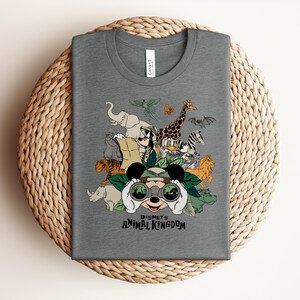 Puede incluir: Una camiseta gris jaspeado de cuello redondo con un estampado gr&aacute;fico de Mickey Mouse mirando a trav&eacute;s de binoculares rodeado de varios animales y personajes de Disney, con el texto "Disney's ANIMAL KINGDOM" impreso en negro bajo la ilustraci&oacute;n.