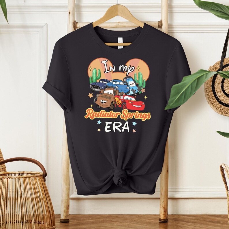 K&ouml;nnte beinhalten: Schwarzes T-Shirt mit einer bunten Grafik von Zeichentrickautos und dem Text "In my Radiator Springs ERA". Das Design umfasst Kakteen und Sterne. Das T-Shirt h&auml;ngt an einem Holzb&uuml;gel.