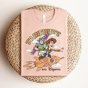 May include: A light peach-colored t-shirt featuring a graphic print of Buzz Lightyear and Woody riding a rocket. The text "TO INFINITY" appears above the characters, and "AND BEYOND" is printed below the rocket, surrounded by small clouds and stars.