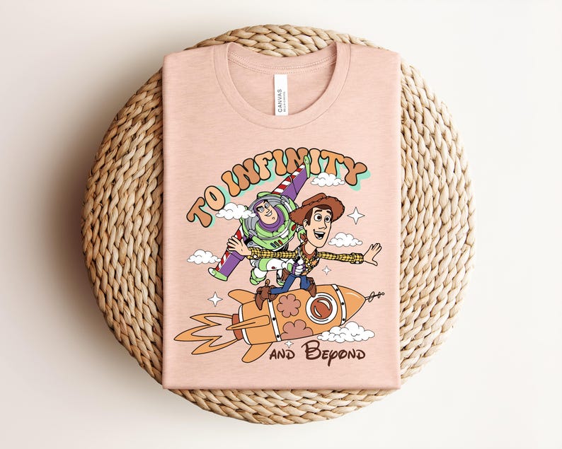 May include: A light peach-colored t-shirt featuring a graphic print of Buzz Lightyear and Woody riding a rocket. The text "TO INFINITY" appears above the characters, and "AND BEYOND" is printed below the rocket, surrounded by small clouds and stars.