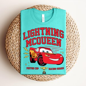 May include: A turquoise t-shirt featuring the text "LIGHTNING McQUEEN" in red, with a red race car graphic. The car has the number 95 on the side and is part of the Piston Cup Racing Series. The shirt is folded on a woven mat.