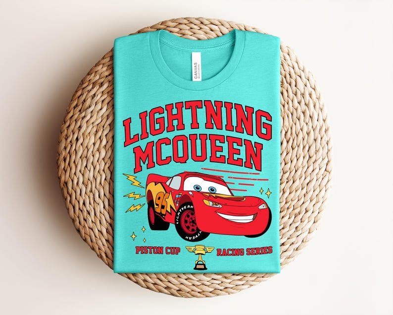 May include: A turquoise t-shirt featuring the text "LIGHTNING McQUEEN" in red, with a red race car graphic. The car has the number 95 on the side and is part of the Piston Cup Racing Series. The shirt is folded on a woven mat.