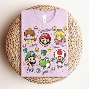 May include: A light purple t-shirt featuring cartoon illustrations of Super Mario characters, including Mario, Luigi, Princess Peach, and Yoshi. The words "Super Mario" and character names are printed on the shirt.