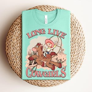 May include: A light mint green t-shirt featuring a graphic print of a cartoon cowgirl riding a brown horse in a desert landscape with cacti. The text "LONG LIVE COWGIRLS" is printed in pink with a dark outline above and below the central illustration.
