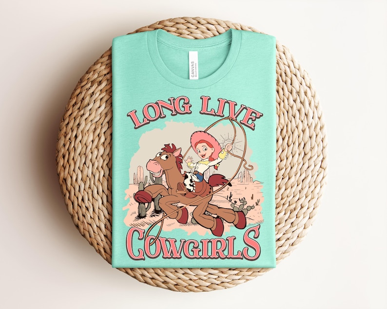 May include: A light mint green t-shirt featuring a graphic print of a cartoon cowgirl riding a brown horse in a desert landscape with cacti. The text "LONG LIVE COWGIRLS" is printed in pink with a dark outline above and below the central illustration.