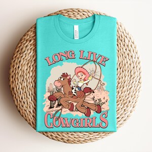 May include: A turquoise t-shirt featuring a graphic print of a cartoon cowgirl riding a brown horse in a desert landscape. The text "LONG LIVE COWGIRLS" is printed in pink above and below the illustration, which includes cacti and a lasso.