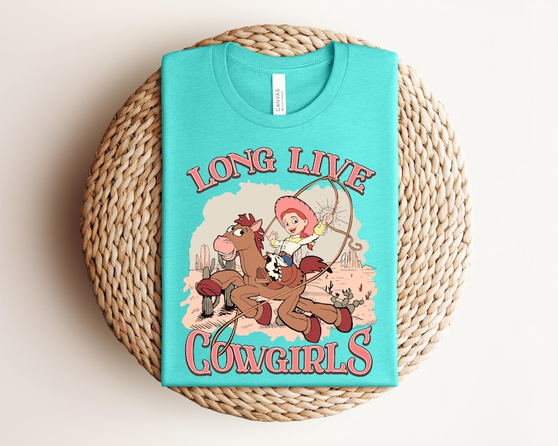May include: A turquoise t-shirt featuring a graphic print of a cartoon cowgirl riding a brown horse in a desert landscape. The text "LONG LIVE COWGIRLS" is printed in pink above and below the illustration, which includes cacti and a lasso.