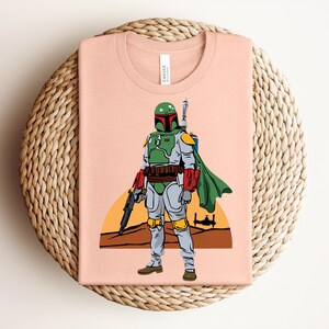 May include: A light peach-colored t-shirt featuring a graphic of a character in green, gray, and red armor, with a green cape and a blaster. The design includes a desert landscape with a setting sun and a spaceship. The shirt is folded on a woven surface.