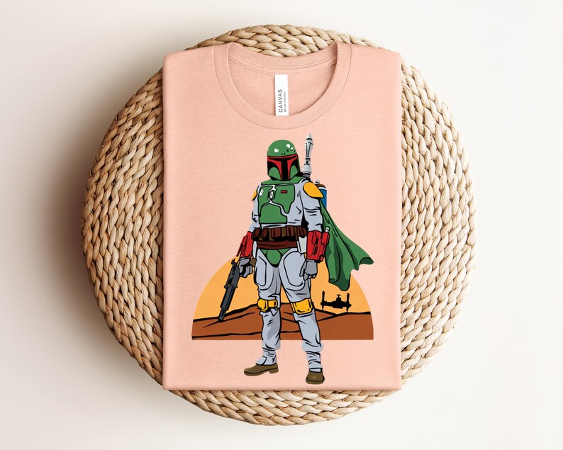 May include: A light peach-colored t-shirt featuring a graphic of a character in green, gray, and red armor, with a green cape and a blaster. The design includes a desert landscape with a setting sun and a spaceship. The shirt is folded on a woven surface.