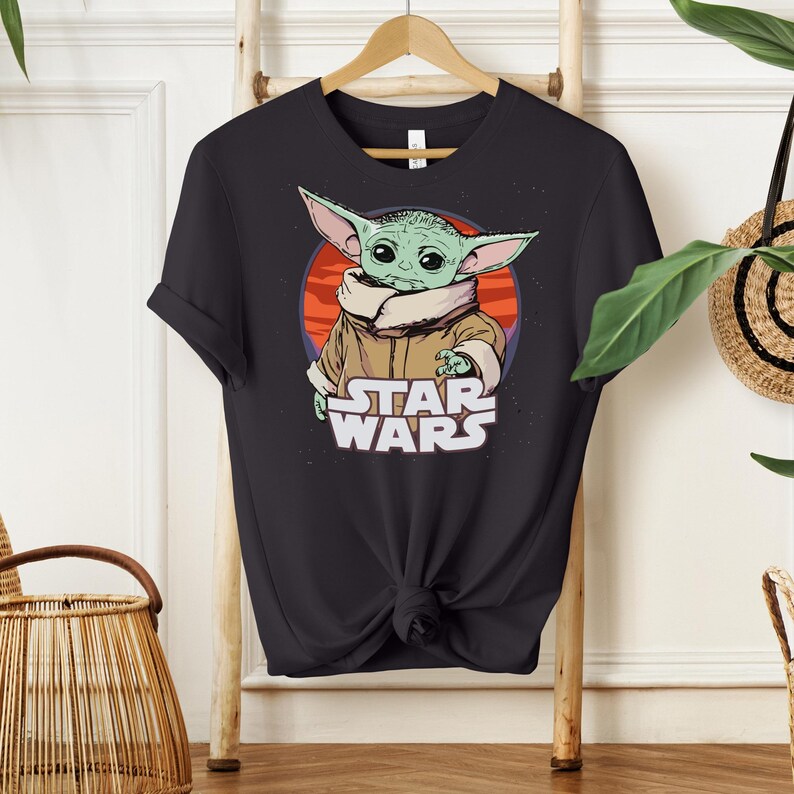 May include: Black t-shirt featuring a graphic of Grogu, also known as Baby Yoda, from Star Wars. The design includes Grogu's image against a red and orange background, with the words "STAR WARS" in white.