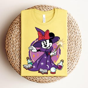 May include: Yellow t-shirt featuring a cartoon Minnie Mouse dressed as a witch, holding a wand with a starburst. She wears a purple dress and hat with a red bow. The design includes potion bottles, books, and a crystal ball.