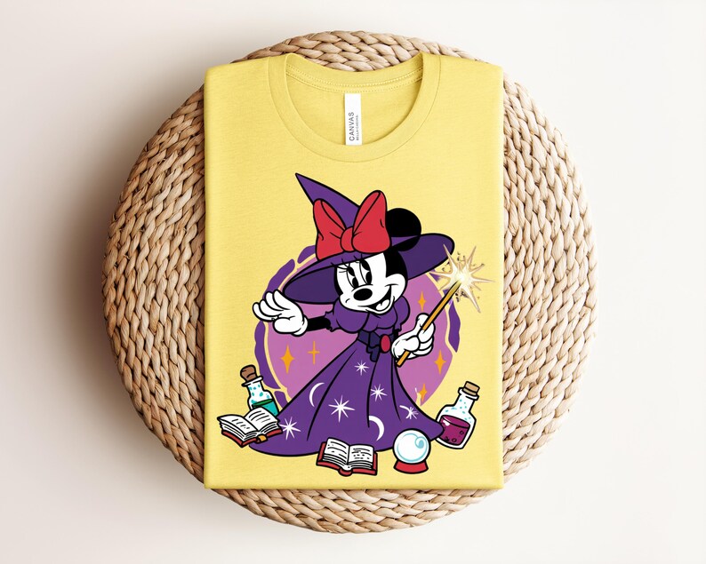 May include: Yellow t-shirt featuring a cartoon Minnie Mouse dressed as a witch, holding a wand with a starburst. She wears a purple dress and hat with a red bow. The design includes potion bottles, books, and a crystal ball.