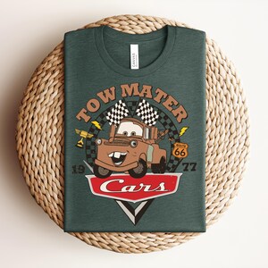 May include: A dark green t-shirt featuring a graphic of the character Tow Mater from the movie Cars. The design includes the text Tow Mater, a checkered flag, a Route 66 sign, a trophy, and the Cars logo with the numbers 19 and 77 on the sides.