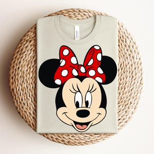 May include: A light beige t-shirt featuring a graphic of Minnie Mouse's face. Minnie's face is black and white with a red bow with white polka dots. The shirt is folded on a woven, circular surface.