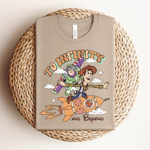 May include: A tan t-shirt featuring a graphic of Buzz Lightyear and Woody from Toy Story. Woody rides an orange rocket while Buzz flies above. The text reads TO INFINITY AND BEYOND in a retro font, surrounded by small white clouds and stars.