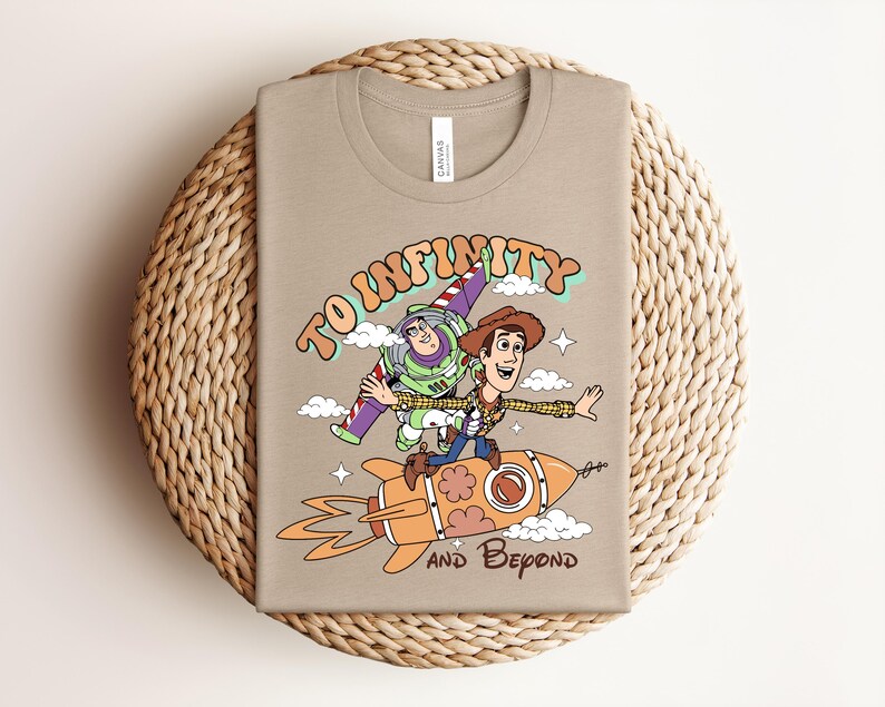 May include: A tan t-shirt featuring a graphic of Buzz Lightyear and Woody from Toy Story. Woody rides an orange rocket while Buzz flies above. The text reads TO INFINITY AND BEYOND in a retro font, surrounded by small white clouds and stars.