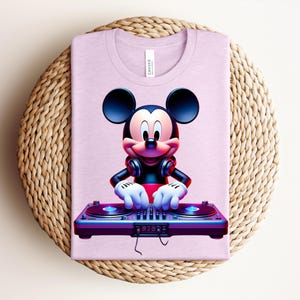 May include: A light purple t-shirt featuring a graphic of Mickey Mouse as a DJ. Mickey is wearing headphones and has his hands on a DJ mixer. The shirt is folded on a woven, circular placemat.