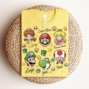 May include: Yellow t-shirt featuring cartoon illustrations of Super Mario characters, including Mario, Luigi, Princess Peach, and Yoshi. The text "Super Mario" and character names are also printed on the shirt.