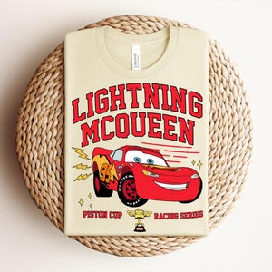 May include: A beige t-shirt featuring a graphic of Lightning McQueen, a red race car with the number 95. The text "LIGHTNING McQUEEN" is in red above the car, with "PISTON CUP RACING SERIES" below. The shirt is folded on a woven mat.