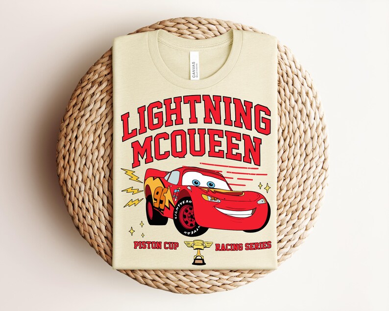 May include: A beige t-shirt featuring a graphic of Lightning McQueen, a red race car with the number 95. The text "LIGHTNING McQUEEN" is in red above the car, with "PISTON CUP RACING SERIES" below. The shirt is folded on a woven mat.