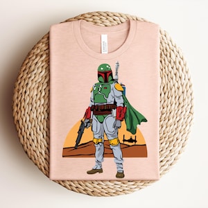 May include: A light pink t-shirt featuring a graphic of a character in green and gray armor, holding a blaster. The character is set against a desert landscape with a setting sun and a spaceship silhouette. The shirt has a tag that says "Canvas".