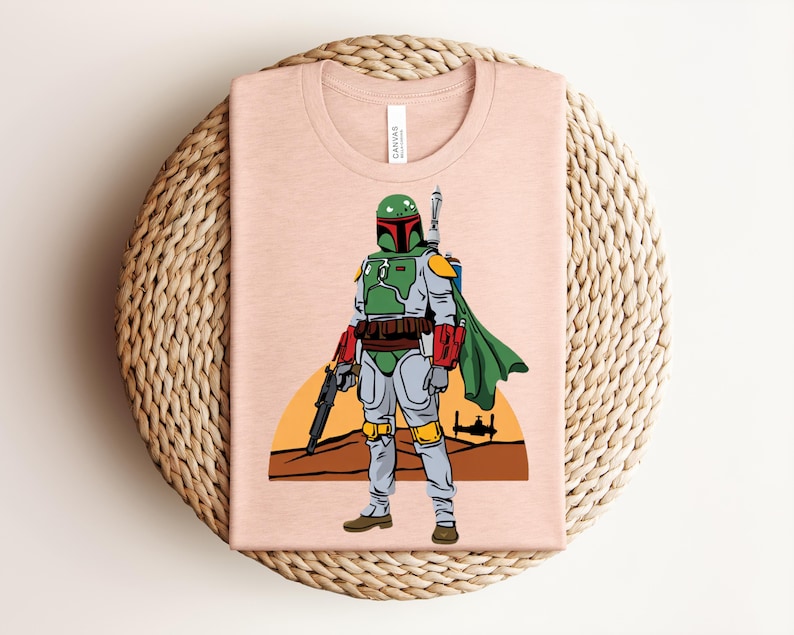 May include: A light pink t-shirt featuring a graphic of a character in green and gray armor, holding a blaster. The character is set against a desert landscape with a setting sun and a spaceship silhouette. The shirt has a tag that says "Canvas".