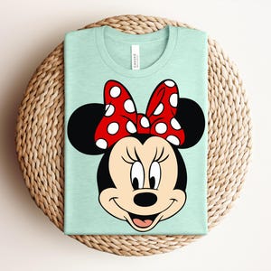 May include: A light green t-shirt featuring a graphic of Minnie Mouse's face. Minnie's face is black with a red bow with white polka dots. The shirt is folded and placed on a woven, circular surface.