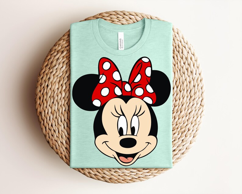 May include: A light green t-shirt featuring a graphic of Minnie Mouse's face. Minnie's face is black with a red bow with white polka dots. The shirt is folded and placed on a woven, circular surface.