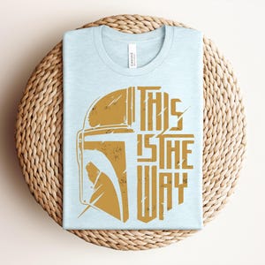 May include: A light blue t-shirt featuring a distressed gold graphic of a helmet on the left side and the text "THIS IS THE WAY" in a bold, stylized font on the right side. The design has a weathered, vintage appearance on the fabric.