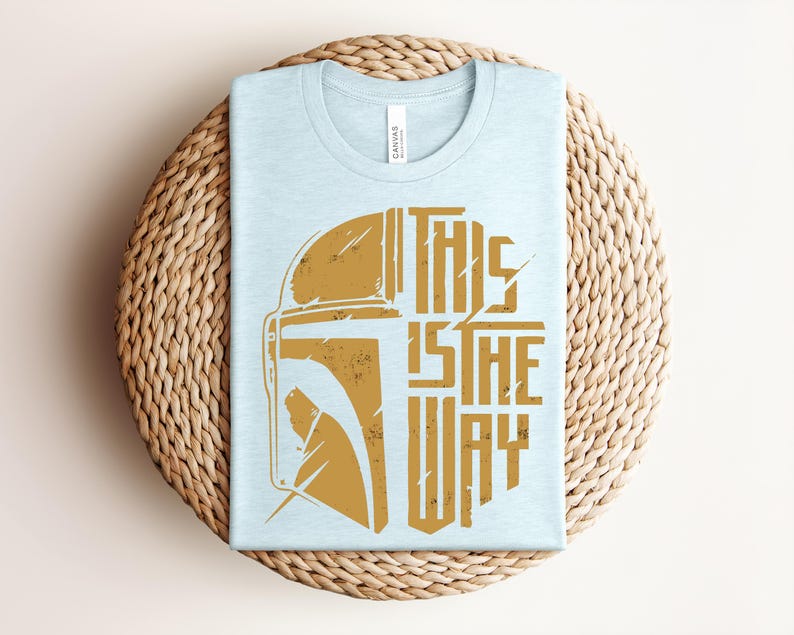 May include: A light blue t-shirt featuring a distressed gold graphic of a helmet on the left side and the text "THIS IS THE WAY" in a bold, stylized font on the right side. The design has a weathered, vintage appearance on the fabric.