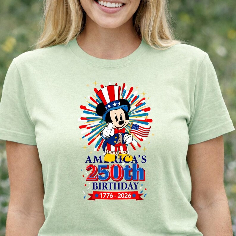 May include: A light green t-shirt featuring a graphic of Mickey Mouse in patriotic attire, holding an American flag. The design celebrates "America's 250th Birthday" with text and dates 1776-2026.