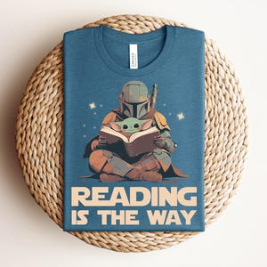 May include: A blue crew neck t-shirt featuring a graphic of a helmeted character in armor sitting cross-legged and reading a book with a small green creature. The text "READING IS THE WAY" is printed in large beige letters at the bottom of the shirt.
