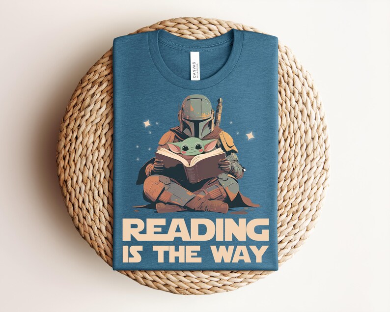 May include: A blue crew neck t-shirt featuring a graphic of a helmeted character in armor sitting cross-legged and reading a book with a small green creature. The text "READING IS THE WAY" is printed in large beige letters at the bottom of the shirt.