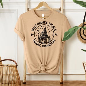 May include: Tan t-shirt with a black graphic of Cinderella's castle inside a circle. The text reads "WALT DISNEY WORLD MAGIC KINGDOM 1971". The shirt is hanging on a wooden hanger.