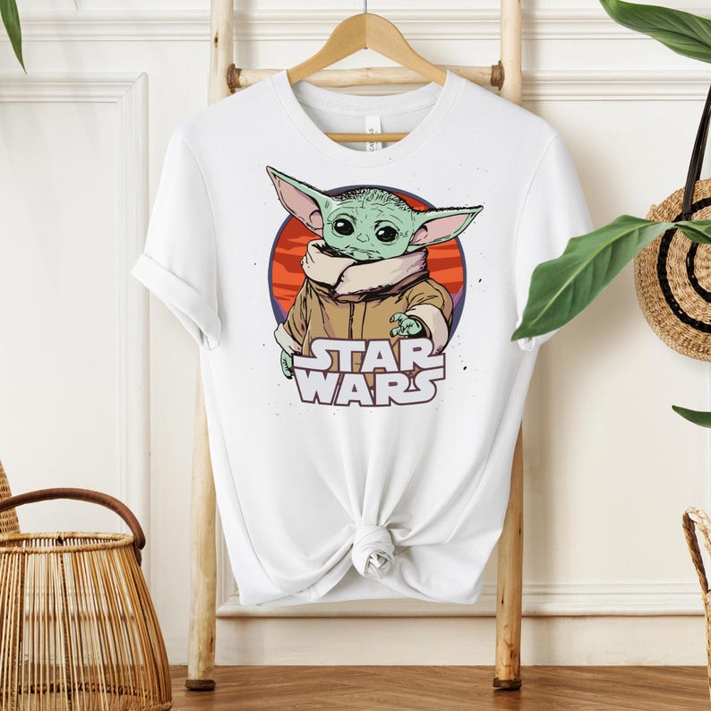 May include: White t-shirt featuring a graphic of a green alien character with large ears, wearing a brown coat, set against a red and orange circle. The words "STAR WARS" are printed below the character.