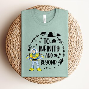Puede incluir: Una camiseta verde claro con un gr&aacute;fico de un personaje de guardi&aacute;n espacial con traje blanco, azul y amarillo. El texto "TO INFINITY AND BEYOND" aparece en negro, rodeado de ilustraciones negras de flechas, estrellas, nubes y un planeta.
