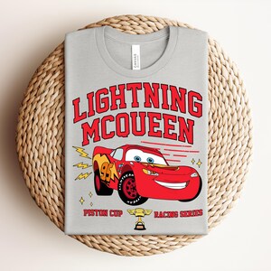 May include: A light gray t-shirt featuring a graphic of Lightning McQueen, a red race car, with the text "LIGHTNING McQUEEN" in red. The shirt also includes the text "Piston Cup Racing Series".