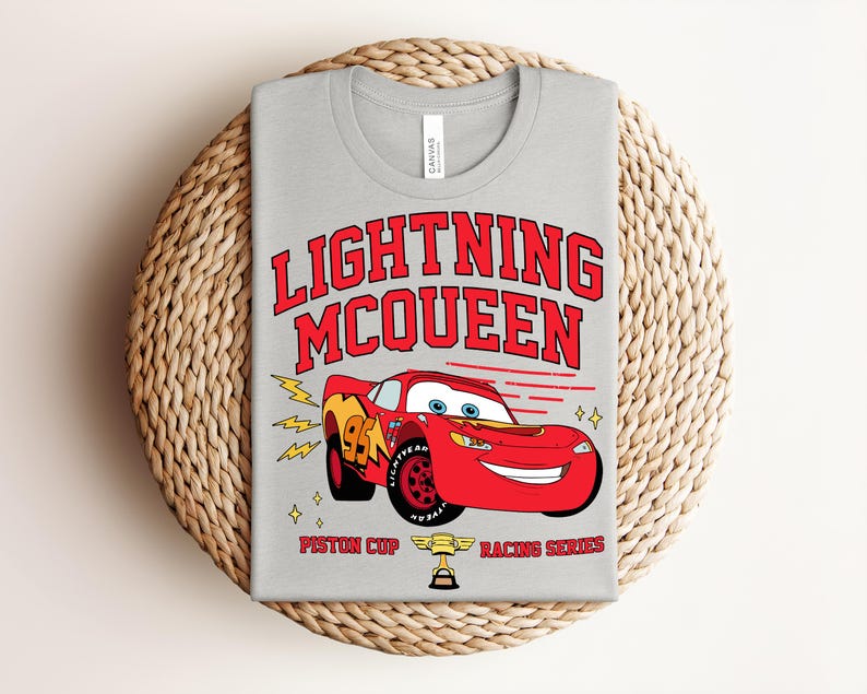 May include: A light gray t-shirt featuring a graphic of Lightning McQueen, a red race car, with the text "LIGHTNING McQUEEN" in red. The shirt also includes the text "Piston Cup Racing Series".