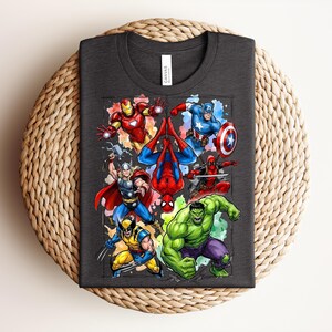 May include: Dark grey t-shirt with a vibrant graphic of Marvel superheroes, such as Iron Man, Spider-Man, Captain America, and the Hulk. The tee shirt is folded on a woven surface. The shirt is made of a soft material.