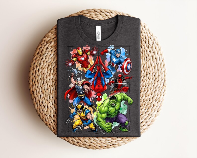May include: Dark grey t-shirt with a vibrant graphic of Marvel superheroes, such as Iron Man, Spider-Man, Captain America, and the Hulk. The tee shirt is folded on a woven surface. The shirt is made of a soft material.