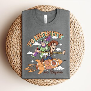May include: A dark gray t-shirt featuring a graphic of Buzz Lightyear and Woody from Toy Story. The design shows them riding an orange rocket with the text TO INFINITY AND BEYOND in a retro font, surrounded by small white clouds and stars.