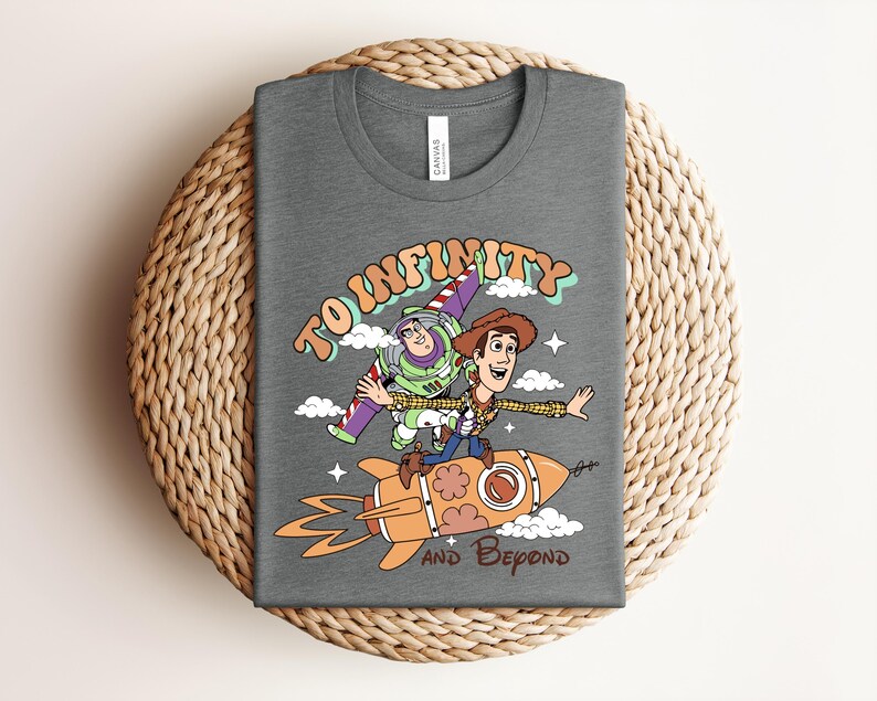 May include: A dark gray t-shirt featuring a graphic of Buzz Lightyear and Woody from Toy Story. The design shows them riding an orange rocket with the text TO INFINITY AND BEYOND in a retro font, surrounded by small white clouds and stars.