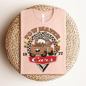 May include: A light peach-colored t-shirt featuring a graphic of the character Tow Mater from the movie Cars. The design includes a checkered flag, a gold trophy, a Route 66 sign, the text Tow Mater, 1977, and the red Cars logo at the bottom.