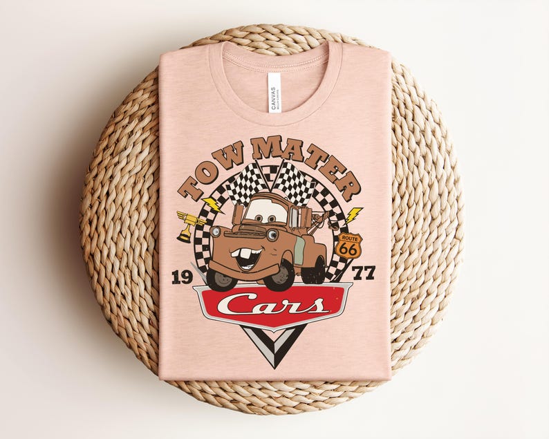 May include: A light peach-colored t-shirt featuring a graphic of the character Tow Mater from the movie Cars. The design includes a checkered flag, a gold trophy, a Route 66 sign, the text Tow Mater, 1977, and the red Cars logo at the bottom.