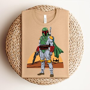 May include: Tan t-shirt featuring a cartoon illustration of a character in green and gray armor, holding a blaster. The character has a green helmet and a green cape. The background shows a sunset and a spaceship.