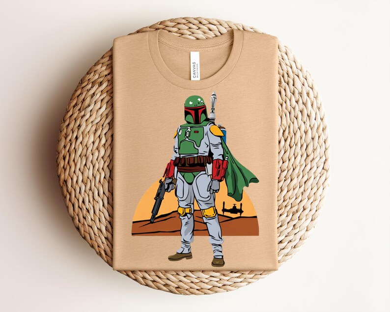 May include: Tan t-shirt featuring a cartoon illustration of a character in green and gray armor, holding a blaster. The character has a green helmet and a green cape. The background shows a sunset and a spaceship.